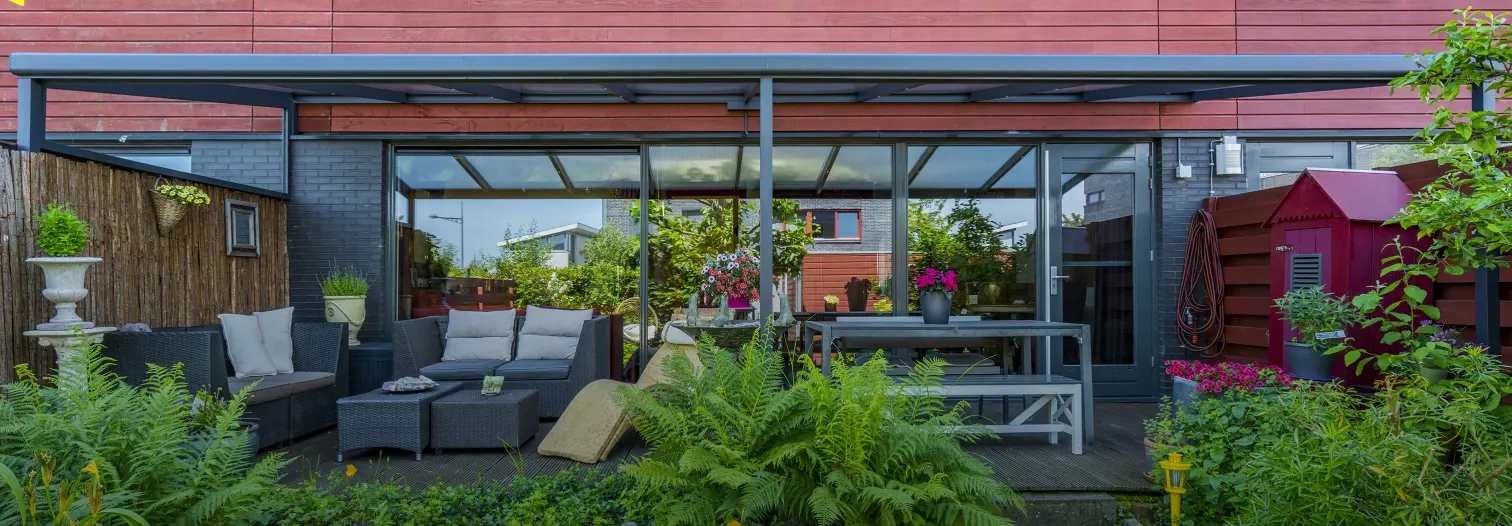 Large covered outdoor patio area with glass walls, modern seating, greenery, and garden elements under a red roof.