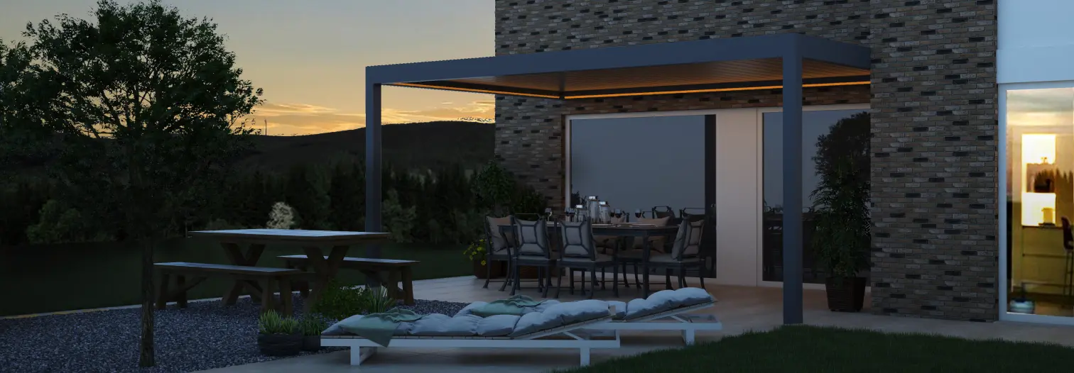 Modern patio scene at dusk with a pergola over a dining table set for a meal. Nearby, lounge chairs face the sunset. A brick wall and tree add warmth