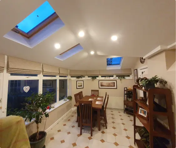 A light-filled conservatory dining area with a wooden table and chairs, tiled floor, skylights in the ceiling, and potted plants along the windows.